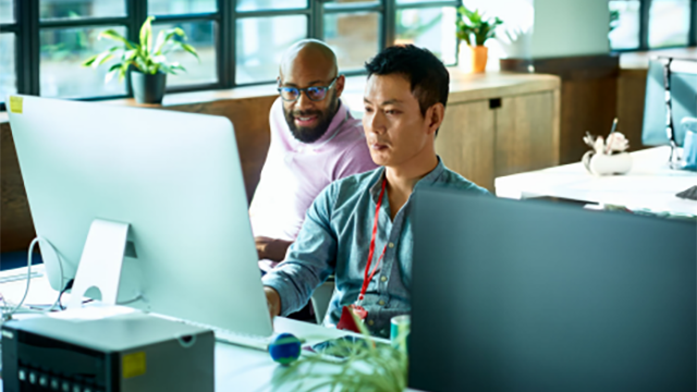 Photograph - Two people sit at a desk in consultation in front of a large monitor.